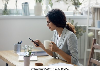 Young Caucasian Woman In Glasses Sit At Desk Drink Coffee Browse Surf Wireless Internet On Modern Cellphone, Calm Female Relax At Workplace Texting Or Messaging, Using Smartphone At Home