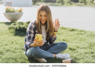 Young Caucasian Woman Having A Video Call Chat With Cellphone, Sitting On The Grass Outside In Park. Happy And Smiling Girl Relax Outdoors. Using Mobile Phone. Distance Learning Online Education.