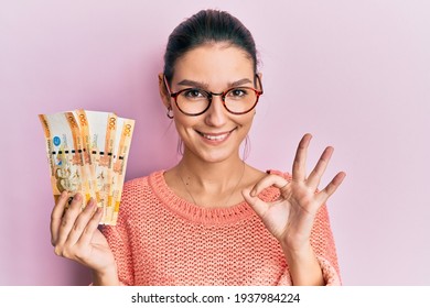 Young caucasian woman holding 500 philippine peso banknotes doing ok sign with fingers, smiling friendly gesturing excellent symbol 
