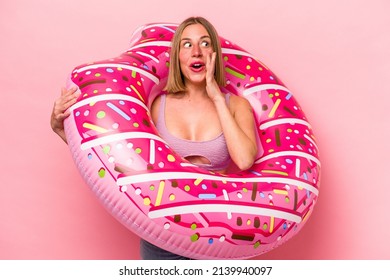 Young caucasian woman holding an air mattress isolated on pink background is saying a secret hot braking news and looking aside