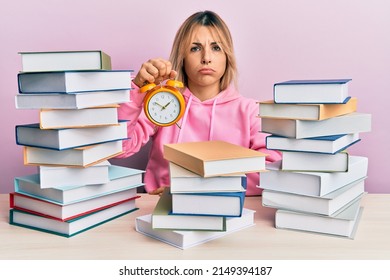 Young caucasian woman holding alarm clock sitting on the table with books depressed and worry for distress, crying angry and afraid. sad expression. 