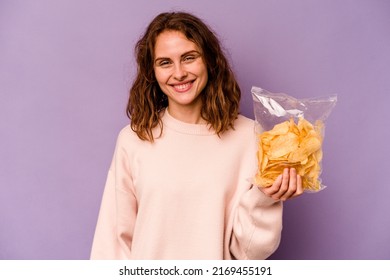 Young Caucasian Woman Holding A Bag Of Chips Isolated On Purple Background Happy, Smiling And Cheerful.