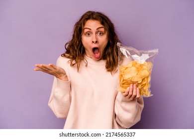 Young Caucasian Woman Holding A Bag Of Chips Isolated On Purple Background Surprised And Shocked.