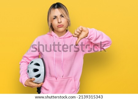 Young caucasian woman holding bike helmet with angry face, negative sign showing dislike with thumbs down, rejection concept 