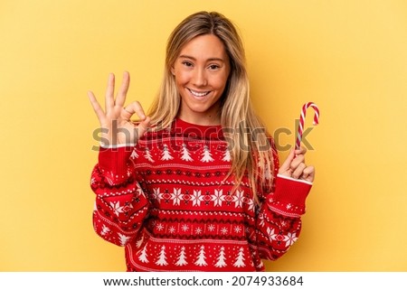 Young caucasian woman holding a christmas stick isolated on yellow background cheerful and confident showing ok gesture.