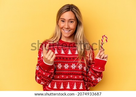 Young caucasian woman holding a christmas stick isolated on yellow background pointing with finger at you as if inviting come closer.