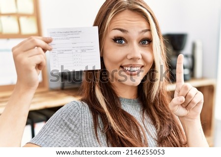 Young caucasian woman holding covid record card surprised with an idea or question pointing finger with happy face, number one 