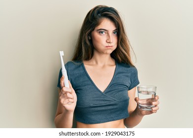 Young caucasian woman holding electric toothbrush and water glass puffing cheeks with funny face. mouth inflated with air, catching air.  - Powered by Shutterstock