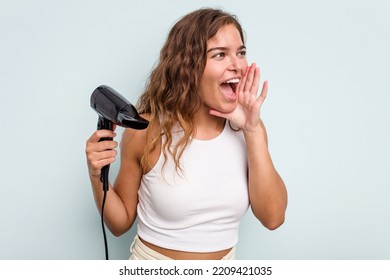 Young Caucasian Woman Holding A Hairdryer Isolated On Blue Background Shouting And Holding Palm Near Opened Mouth.