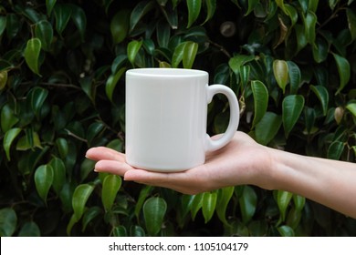 Young Caucasian Woman Holding In Hand White Mockup Mug On Green Tree Foliage Nature Background In Forest. Blank Space For Lettering Artwork Product Branding. Eco Organic Hipster Style.