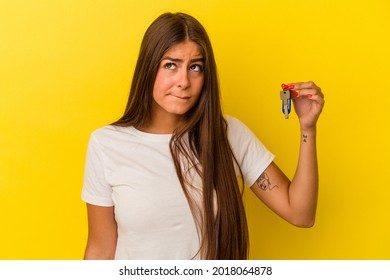 Young caucasian woman holding a home keys isolated on yellow background confused, feels doubtful and unsure.