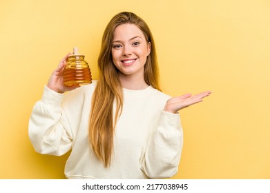 Young Caucasian Woman Holding A Honey Isolated On Yellow Background Showing A Copy Space On A Palm And Holding Another Hand On Waist.