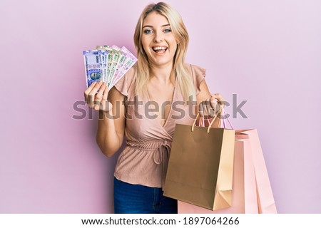 Young caucasian woman holding indian rupee banknotes and shopping bags smiling and laughing hard out loud because funny crazy joke. 