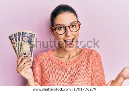 Young caucasian woman holding japanese yen banknotes celebrating achievement with happy smile and winner expression with raised hand 