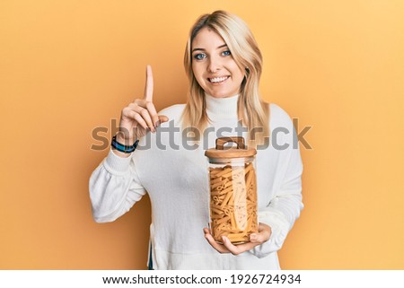 Young caucasian woman holding jar of macaroni pasta smiling with an idea or question pointing finger with happy face, number one 