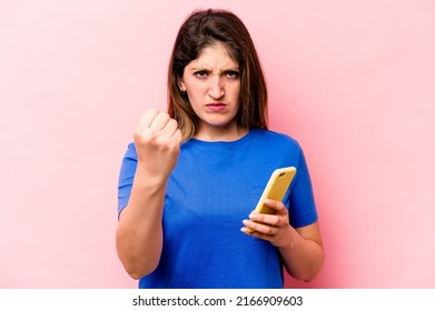 Young Caucasian Woman Holding Mobile Phone Isolated On Pink Background Showing Fist To Camera, Aggressive Facial Expression.