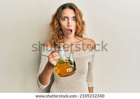 Young caucasian woman holding olive oil can scared and amazed with open mouth for surprise, disbelief face 