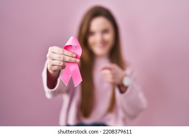 Young caucasian woman holding pink cancer ribbon smiling happy pointing with hand and finger  - Shutterstock ID 2208379611