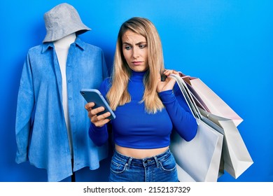 Young Caucasian Woman Holding Shopping Bags And Smartphone Clueless And Confused Expression. Doubt Concept. 