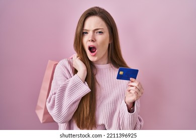 Young Caucasian Woman Holding Shopping Bag And Credit Card Angry And Mad Screaming Frustrated And Furious, Shouting With Anger. Rage And Aggressive Concept. 