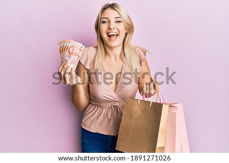 Young caucasian woman holding singapore dollars banknotes and shopping bags smiling and laughing hard out loud because funny crazy joke. 