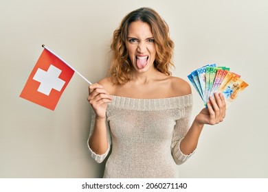 Young caucasian woman holding switzerland flag and franc banknotes sticking tongue out happy with funny expression. 