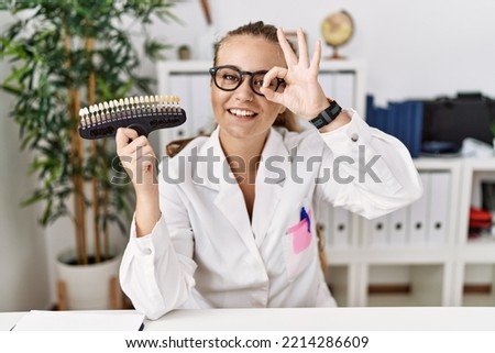 Young caucasian woman holding teeth whitening palette doing ok gesture with hand smiling, eye looking through fingers with happy face. 