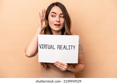 Young caucasian woman holding a virtual reality placard isolated on beige background trying to listening a gossip.