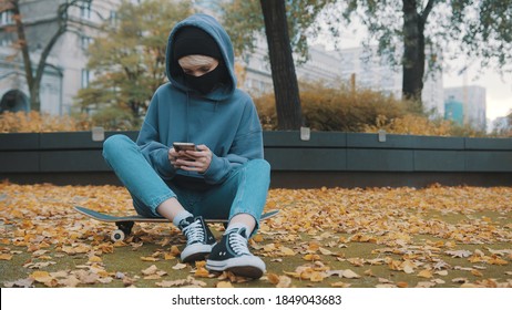 Young caucasian woman with hoodie and facce mask sitting on the skateboard and using smartphone on autumn day. High quality photo - Powered by Shutterstock