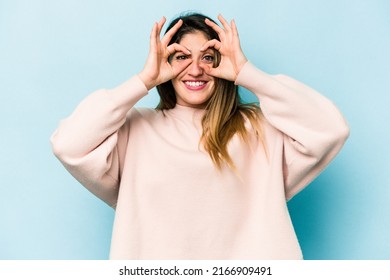 Young caucasian woman isolated on blue background showing okay sign over eyes