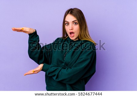 Young caucasian woman isolated on purple background shocked and amazed holding a copy space between hands.