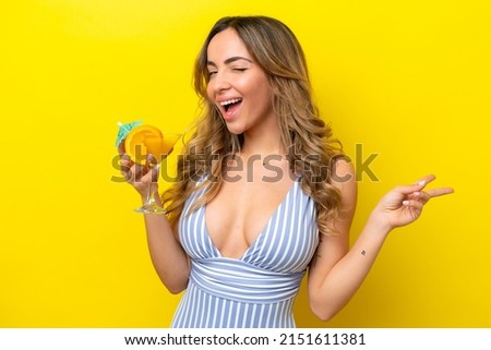 Young caucasian woman isolated on yellow background in swimsuit and holding a cocktail