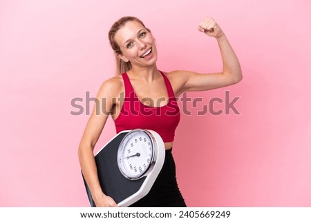 Young caucasian woman isolated on pink background holding a weighing machine and doing strong gesture