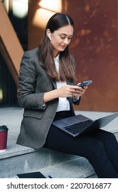 Young Caucasian Woman With Laptop Sitting On The Stairs Holding Phone Reading Good News In A Modern Urban Space In The Financial District. Modern Communication