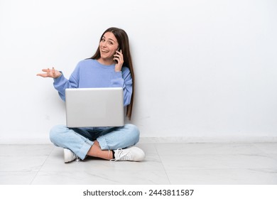 Young caucasian woman with laptop sitting on the floor isolated on white background keeping a conversation with the mobile phone with someone - Powered by Shutterstock