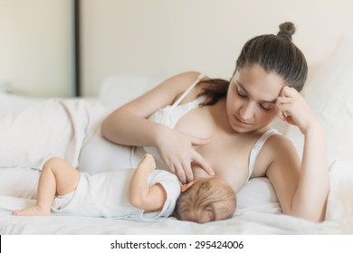 Young Caucasian woman laying in her bedroom on the white cushions, smiling and breastfeed her baby. 