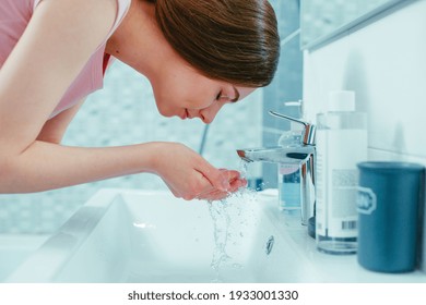 Young Caucasian woman leaning to the sink in the bathroom with her eyes closed and taking handful of water