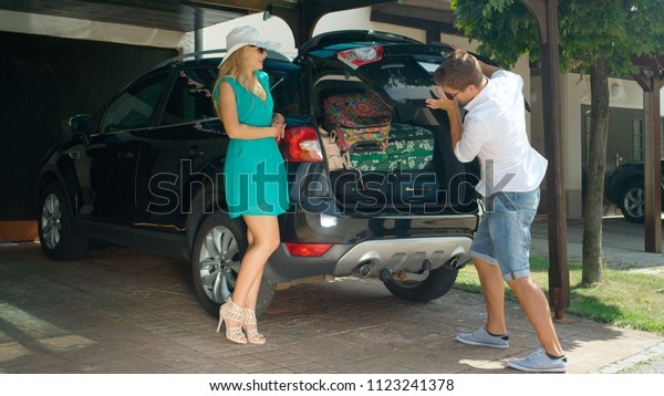 Young Caucasian woman leans on the black car\
while her husband stuffs the trunk with their travel bags. Cheerful\
newlyweds getting their luggage in the trunk of black SUV before\
leaving for honeymoon.