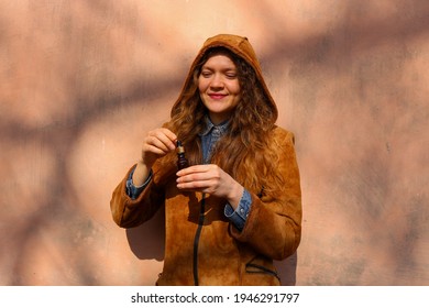 A young caucasian woman with light brown blond curly hair opening a CBD oil bottle with a dripper and smiling. Wearing a brown faux leather jacket with a hood.