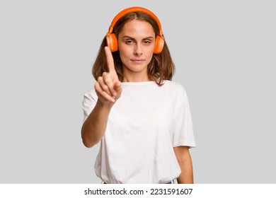 Young caucasian woman listening to music with headphones isolated showing number one with finger.