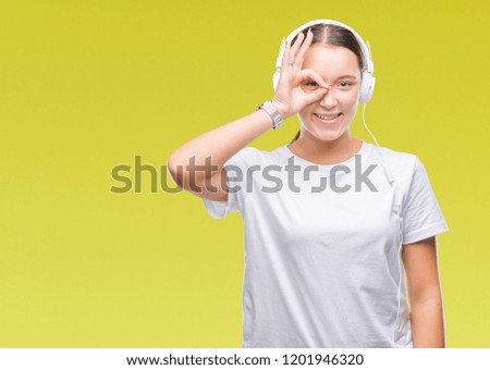 Young caucasian woman listening to music wearing headphones over isolated background doing ok gesture with hand smiling, eye looking through fingers with happy face.