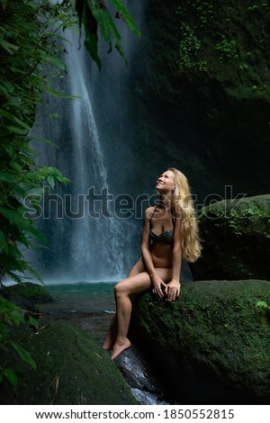 Young Caucasian woman with long blond hair sitting on the rock and enjoying waterfall landscape. Travel lifestyle. Woman wearing bikini. Leke Leke waterfall, Bali, Indonesia