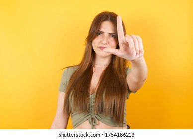 Young caucasian woman with long hair wearing green tshirt standing over yellow isolated background making fun of people with fingers on forehead doing loser gesture mocking and insulting. - Shutterstock ID 1754186213