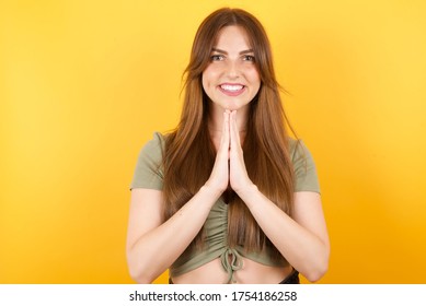 Junge Kaukasierin mit langen Haaren, die grünes T-Shirt tragen, steht auf isoliertem gelbem Hintergrund und betet mit Händen zusammen um Verzeihung lächelnd selbstbewusst.