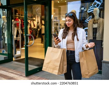 Young Caucasian woman looking at her cell phone in front of a shopping mall window. Bags hanging from her arms. Shopping on a sale day. Black Friday. Plaza Mayor shopping center in Malaga