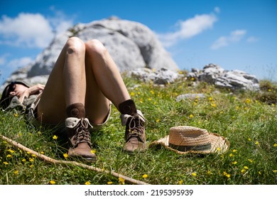 Young Caucasian Woman Lying Down And Taking A Rest In The Warm Sun Near A Wooden Trekking Stick And Wicker Hat In The Grass On A Mountain Peak With Blue Sky And Clouds Above