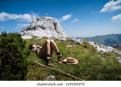 Young Caucasian Woman Lying Down And Taking A Rest In The Warm Sun Near A Wooden Trekking Stick And Wicker Hat In The Grass On A Mountain Peak With Blue Sky And Clouds Above