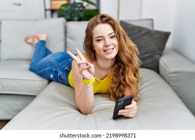 Young Caucasian Woman Lying On The Sofa Using Smartphone Smiling Looking To The Camera Showing Fingers Doing Victory Sign. Number Two. 