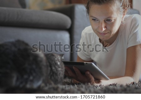 Young caucasian woman lying on the carpet at home, using digital tablet computer and teasing her black pet cat