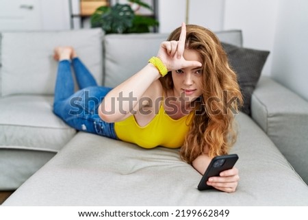 Young caucasian woman lying on the sofa using smartphone making fun of people with fingers on forehead doing loser gesture mocking and insulting. 